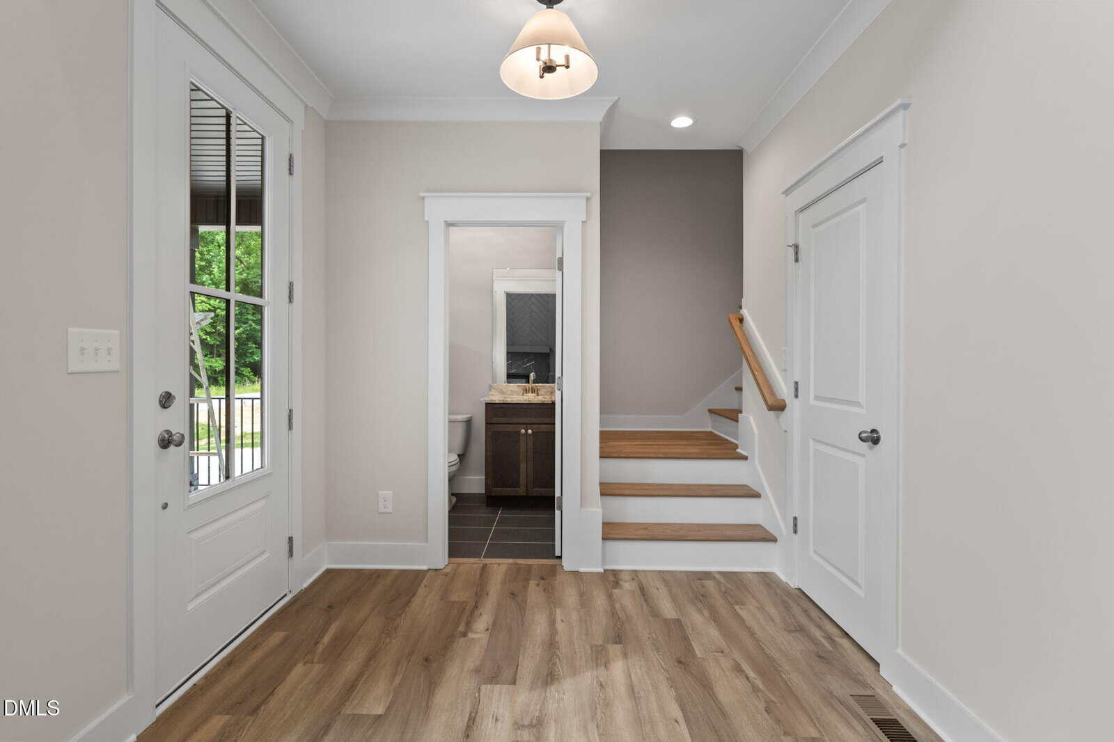 Modern entryway with light wood floors, white walls, a glass door on the left, a small bathroom ahead, and carpeted stairs with a wooden handrail leading up to the right. Ceiling light and natural light brighten the space.