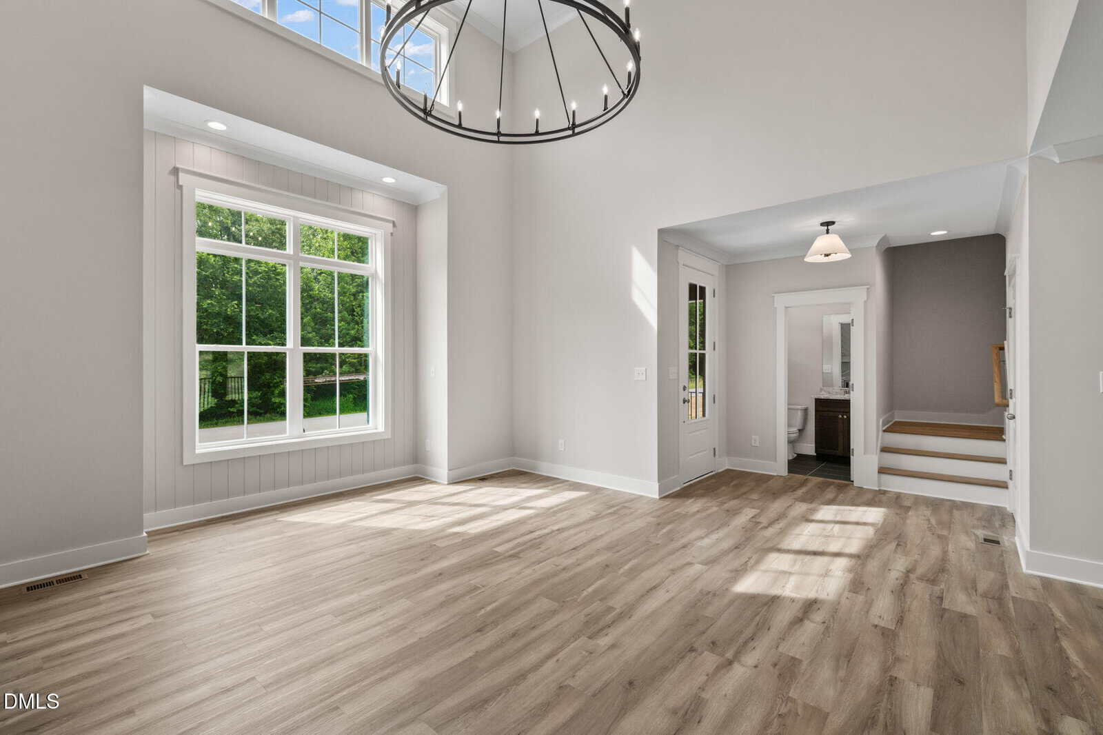 Bright, modern room with large windows, light wood flooring, white walls, high ceiling with a circular chandelier, and an open doorway leading to stairs and a small hallway.