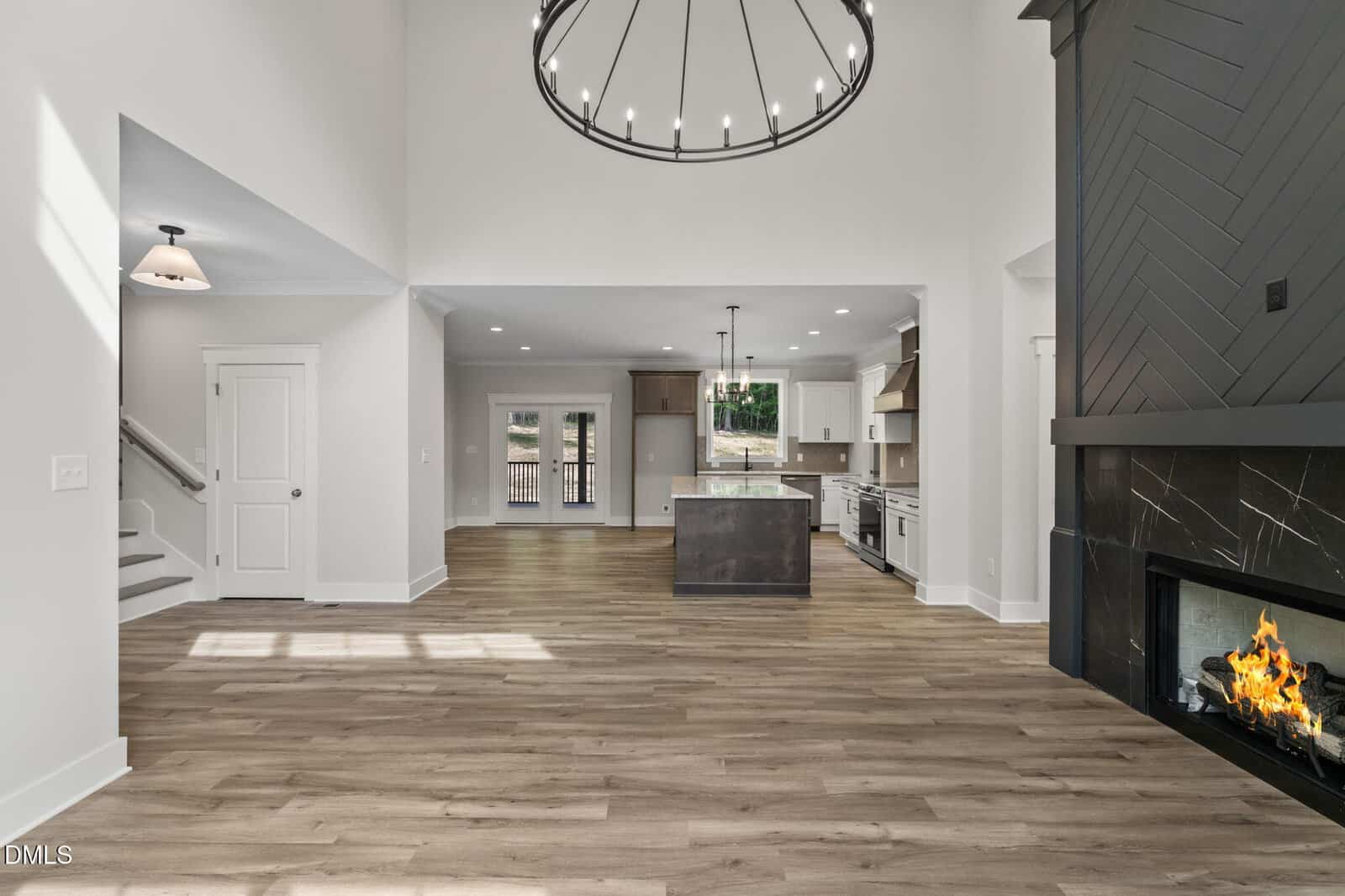 Modern open-concept living area with light wood floors, a black fireplace, large chandelier, kitchen island, and white walls. Stairs and multiple doors are visible, with sunlight streaming through windows.