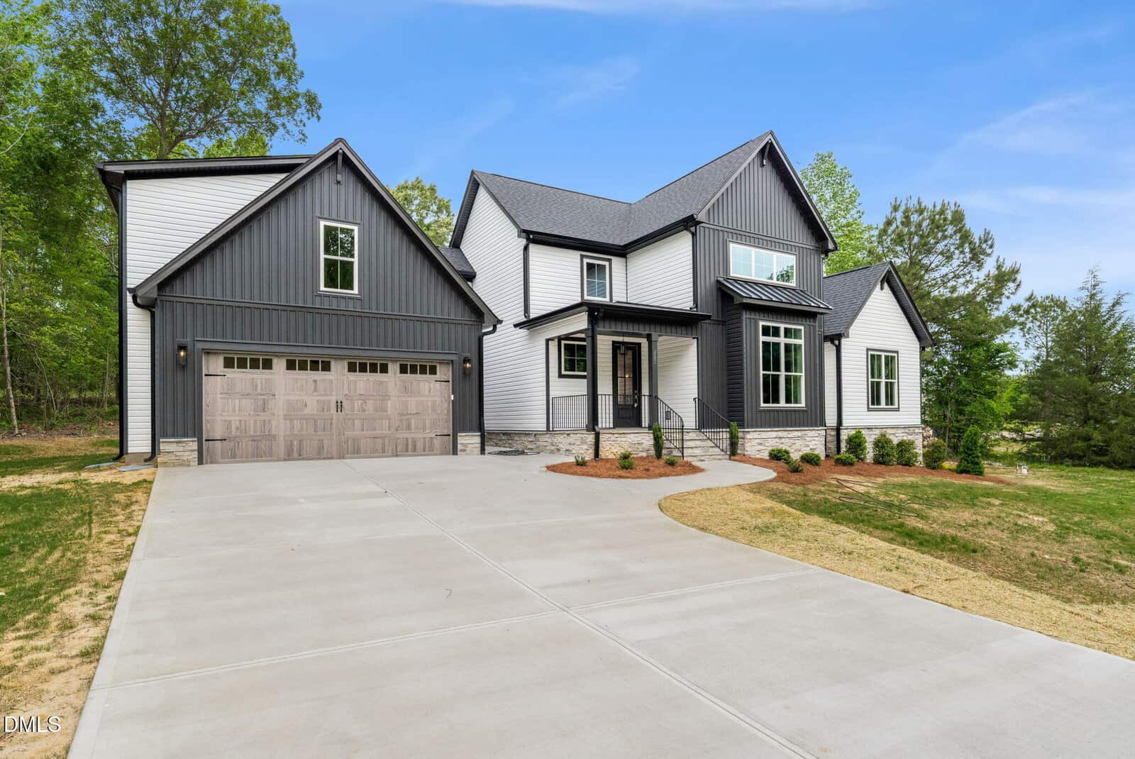 Modern two-story house with black and white exterior, large windows, and a double garage with wooden doors; wide concrete driveway and landscaped yard with small bushes and trees in the background.