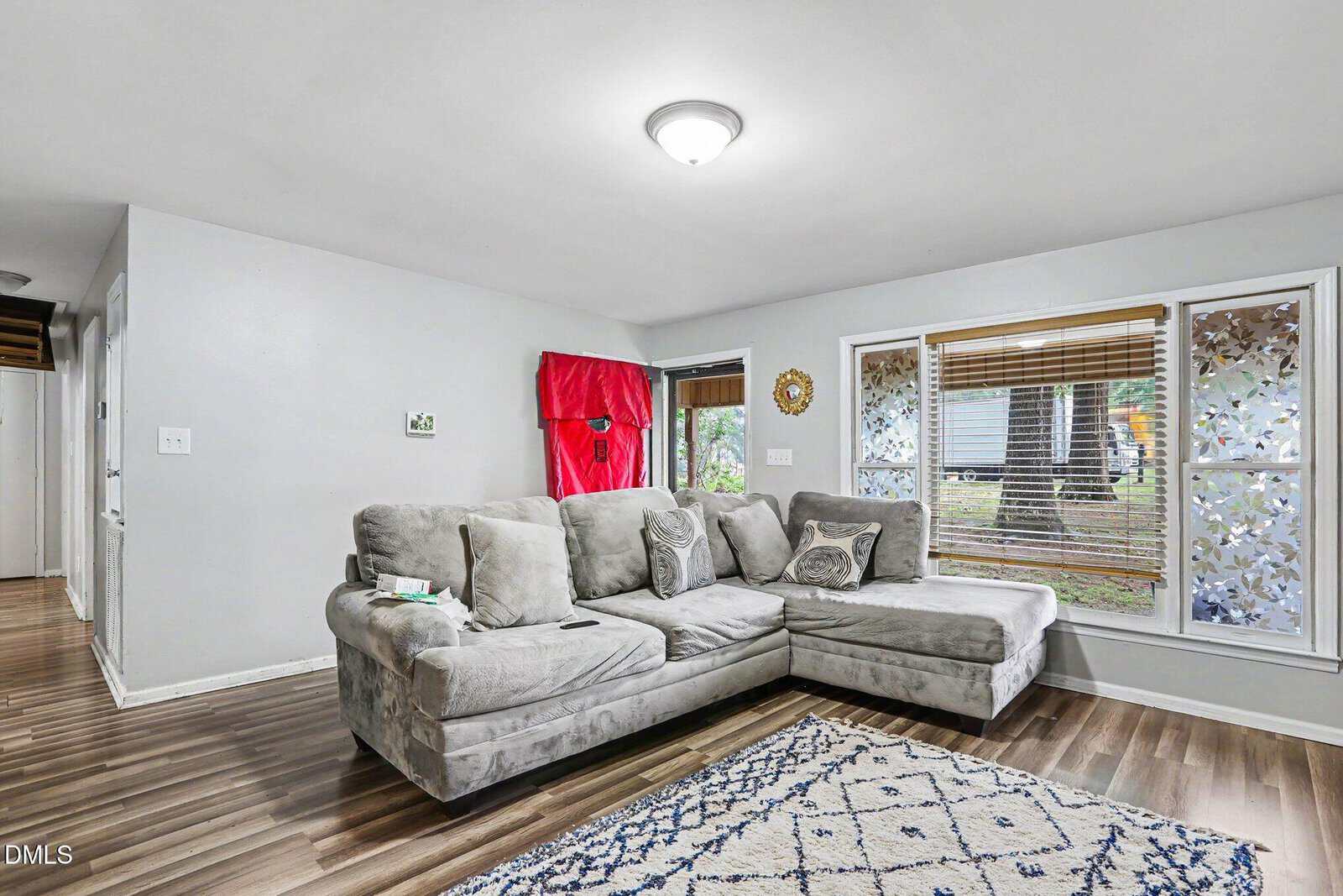 A cozy living room with a gray sectional sofa, patterned rug, and large window with blinds and decorative film. The walls are light gray, and the floor is wood. A red cloth covers an object by the back wall.