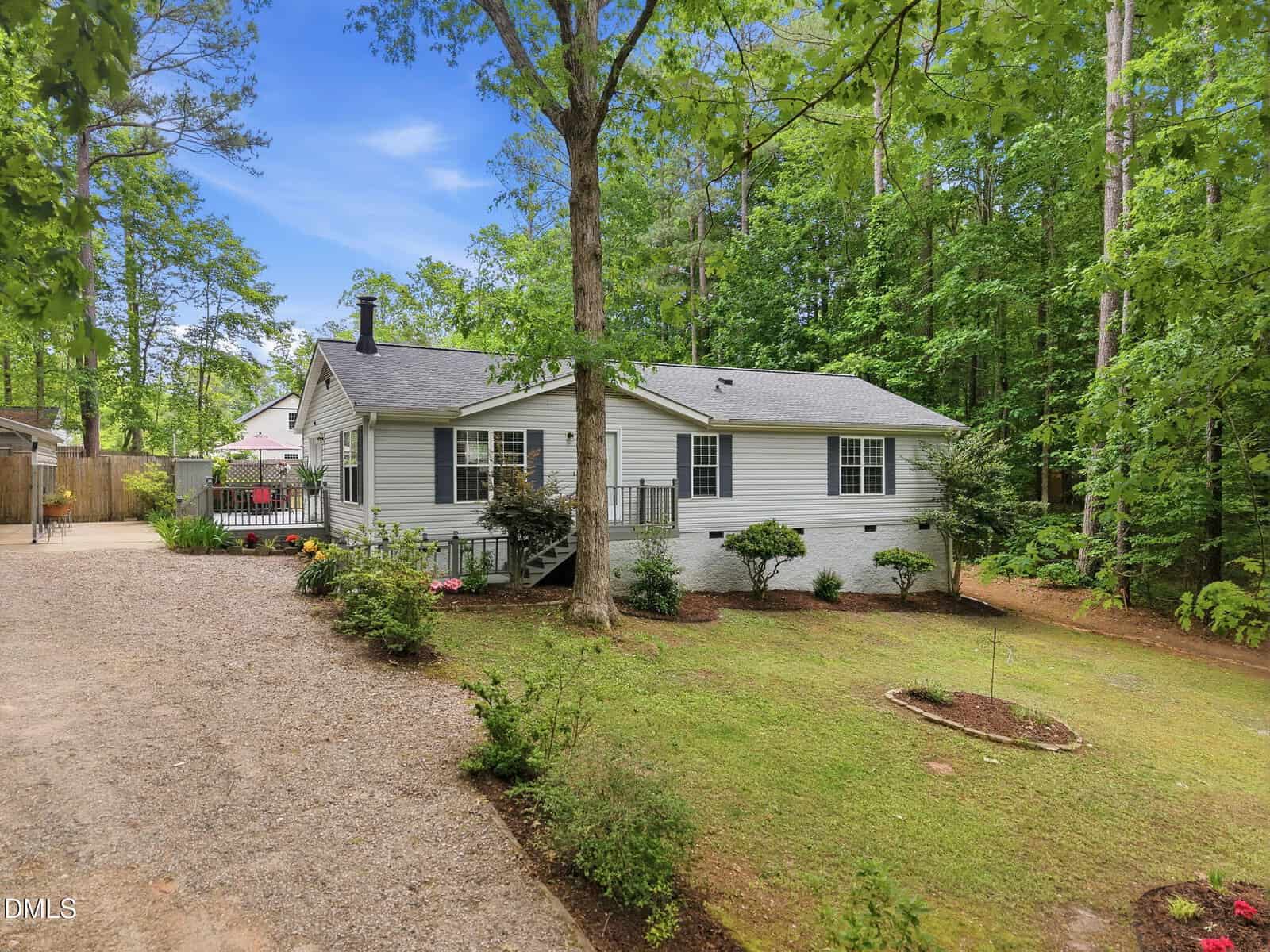 A single-story gray house with white trim sits among tall trees, surrounded by green landscaping. A gravel driveway leads to the house, and a small garden bed decorates the front yard.