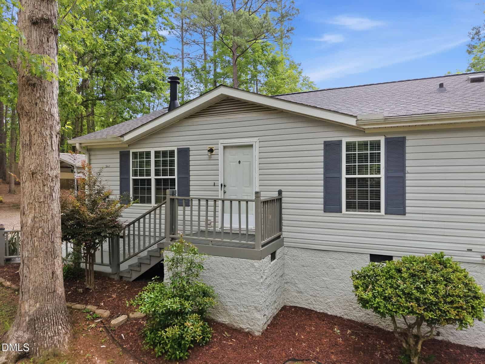 Single-story gray house with white trim, small front porch, and two sets of steps. Surrounded by trees and shrubs, with a fresh mulch bed and a forested background under a blue sky.