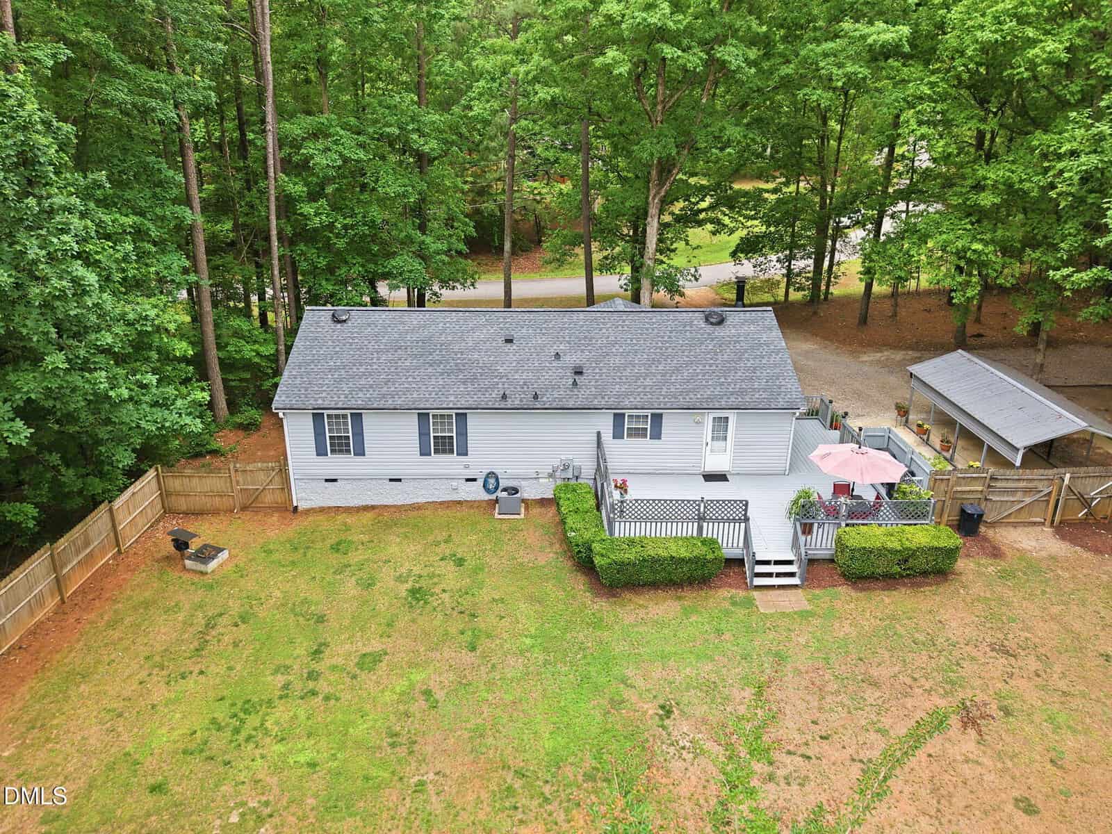 A small, light-gray house with a fenced backyard, surrounded by trees. The yard has a fire pit, deck with patio furniture and an umbrella, and a covered parking area to the right. A road is visible beyond the fence.