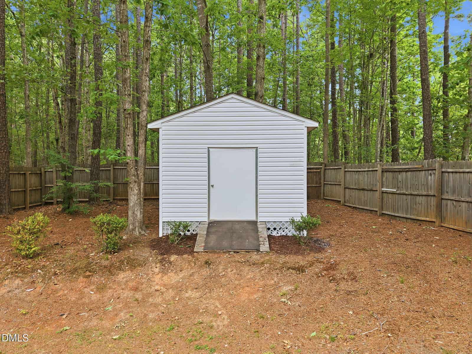A small white shed with a central door stands in a fenced backyard surrounded by tall trees. The shed sits on a patch of bare ground with some green shrubs and pine needles scattered around.