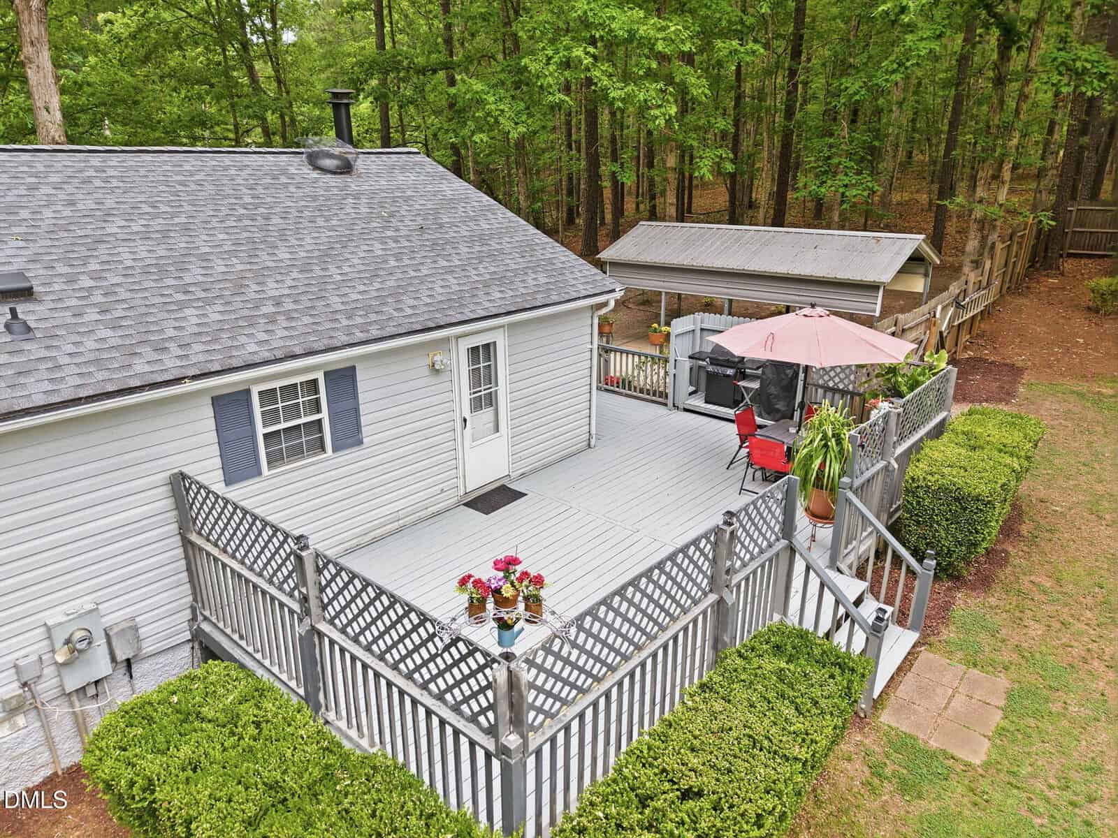 A gray house with a spacious wooden deck featuring potted plants, seating, a red umbrella, and a fenced area. The deck overlooks a green yard and is bordered by trees and a wooded area.