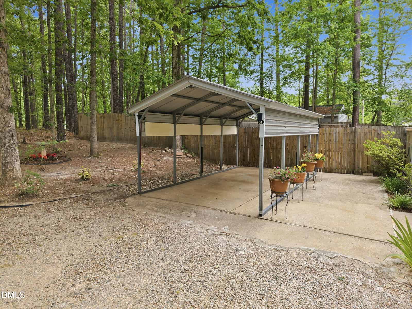 A metal carport with a concrete pad, surrounded by trees and wooden fencing. Potted plants sit on metal stands along the right edge, and a small garden area is on the left.