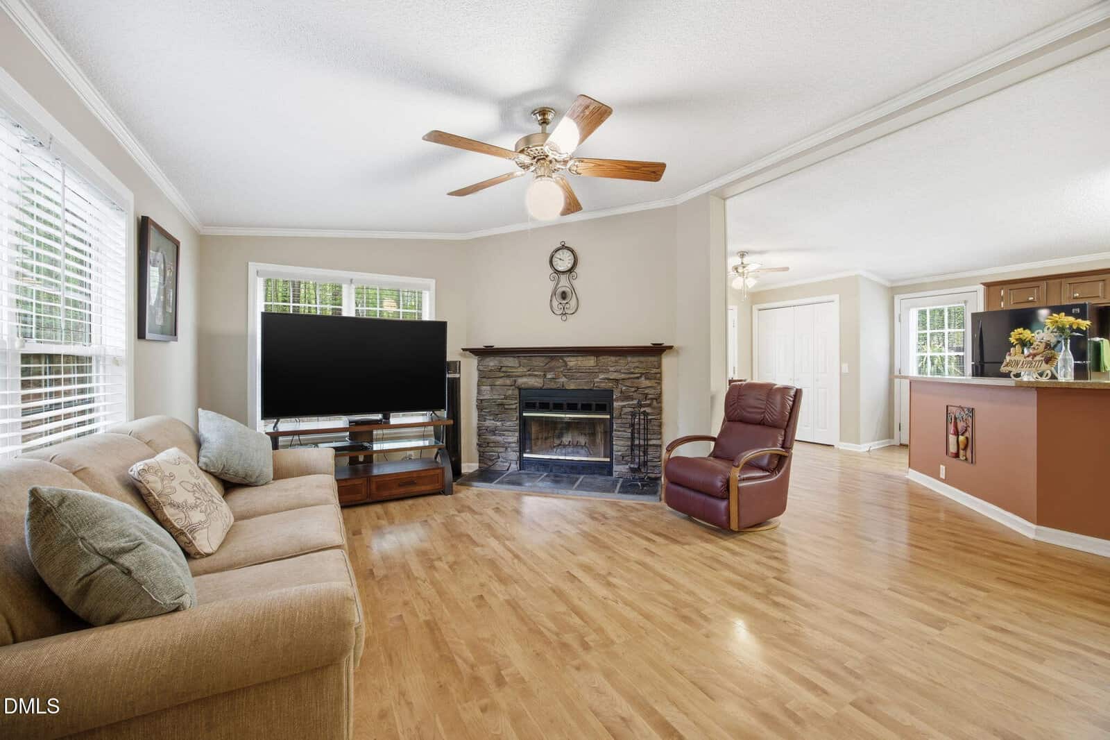 Living room with wood floors, a beige sofa, brown recliner, TV on a stand, and a stone fireplace. Ceiling fan, windows with blinds, and an open view into the kitchen and dining area.