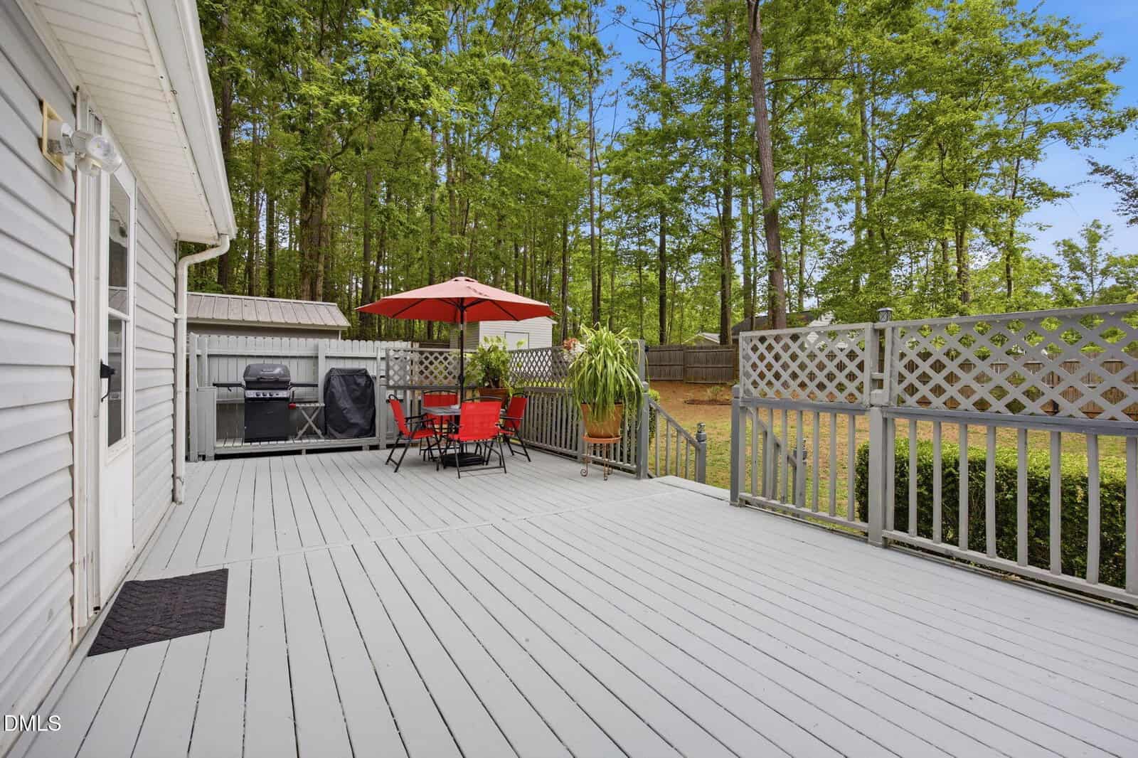 Spacious gray wooden deck with red patio set under a red umbrella, grill, potted plants, and lattice fencing, overlooking a fenced backyard and wooded area.