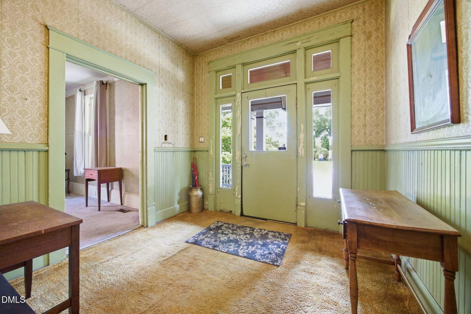 Vintage entryway with pale green wood paneling, patterned wallpaper, and tan carpet. Glass-paneled front door lets in natural light; an area rug and wooden tables sit against the walls. A nearby room is visible through an open doorway.