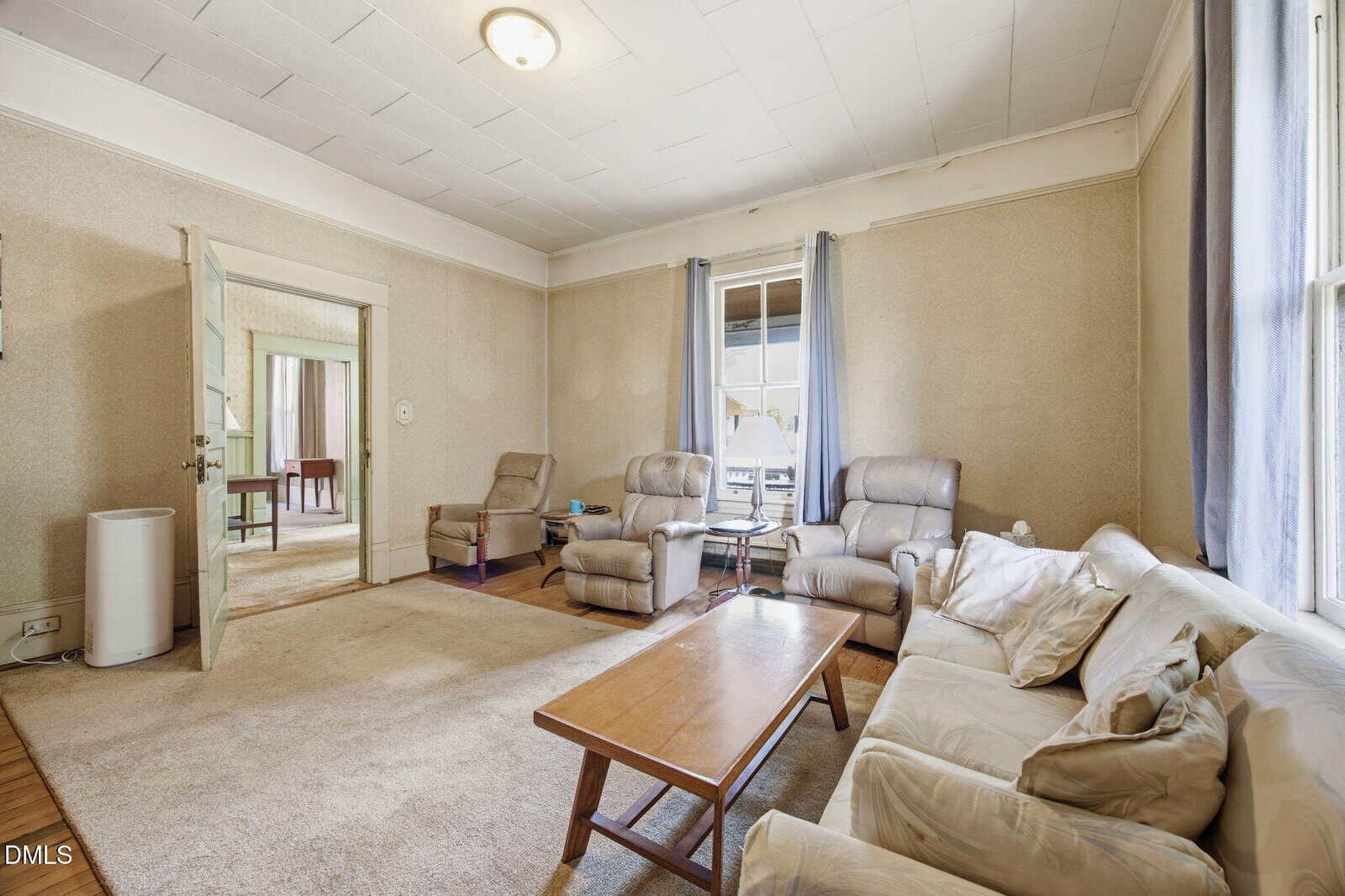 A spacious living room with beige walls, two recliners, a cream sofa, wooden coffee table, and large windows with gray curtains. The room has carpeted and hardwood floors and an open doorway to another room.