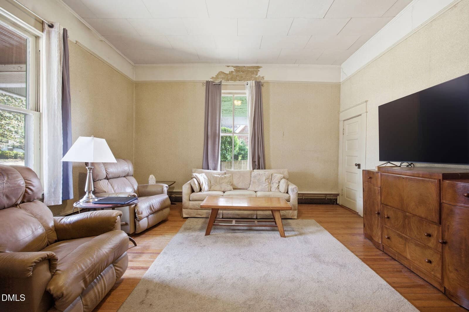 A living room with beige walls and ceiling damage above a window, furnished with two brown recliners, a cream sofa, a wooden coffee table, a rug, a TV on a wooden stand, and two tall windows with curtains.