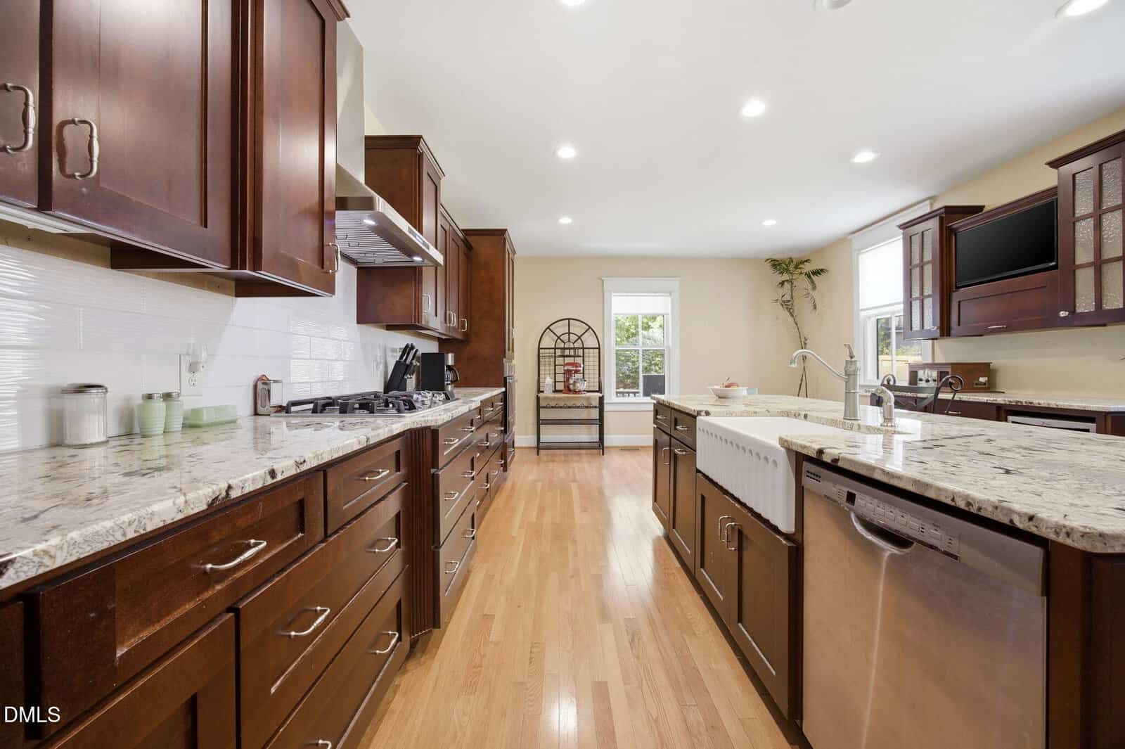 Spacious kitchen with wooden cabinets, marble countertops, stainless steel appliances, farmhouse sink, and a large window allowing natural light to fill the room. Light wood flooring and a decorative rack by the window.