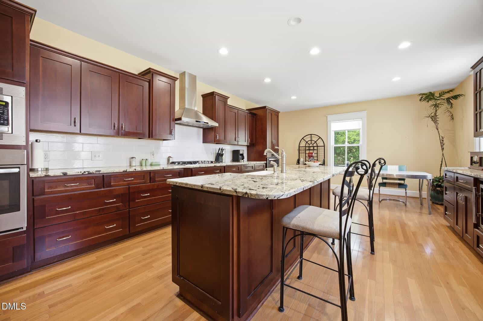 Spacious kitchen with dark wood cabinets, granite island with bar stools, stainless steel appliances, and hardwood floors. Bright window and a small desk with a chair in the background.