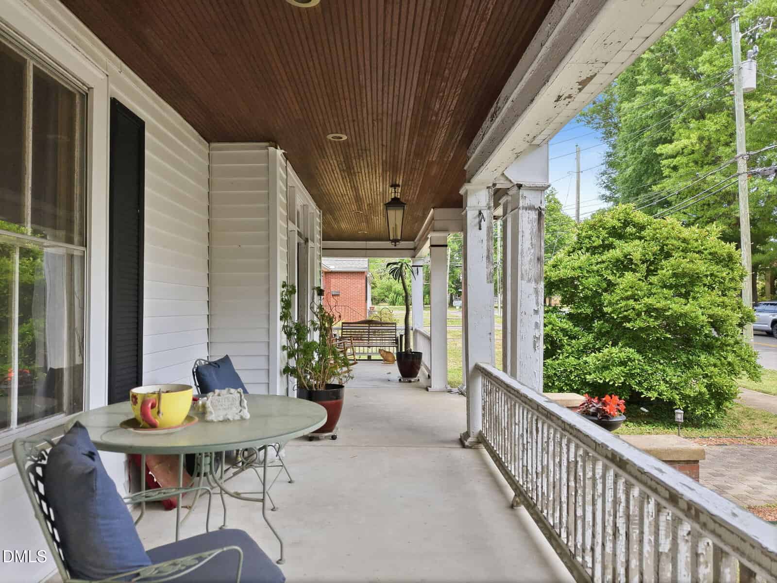 A spacious covered front porch with a wooden ceiling, white railing, potted plants, and a small round table with two chairs. The porch overlooks a green yard and street with trees and power lines.