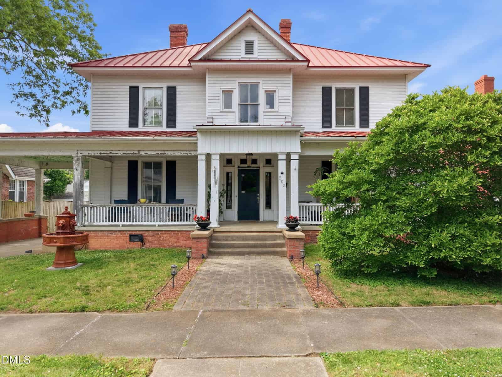 Two-story white house with red metal roof, large covered front porch, black shutters, and columns. Brick steps lead to double front doors. A green bush and fountain are in the yard, with a sidewalk in front.