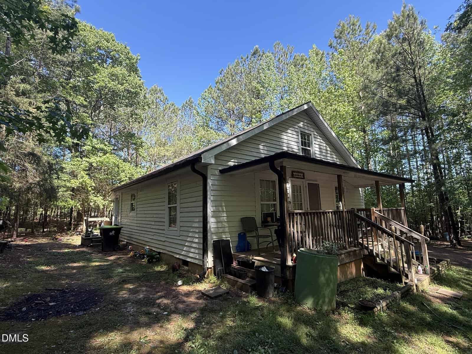 A small white house with a covered front porch sits among tall trees and greenery on a sunny day. Outdoor chairs and miscellaneous items are on the porch and near the house. The yard is shaded and surrounded by forest.