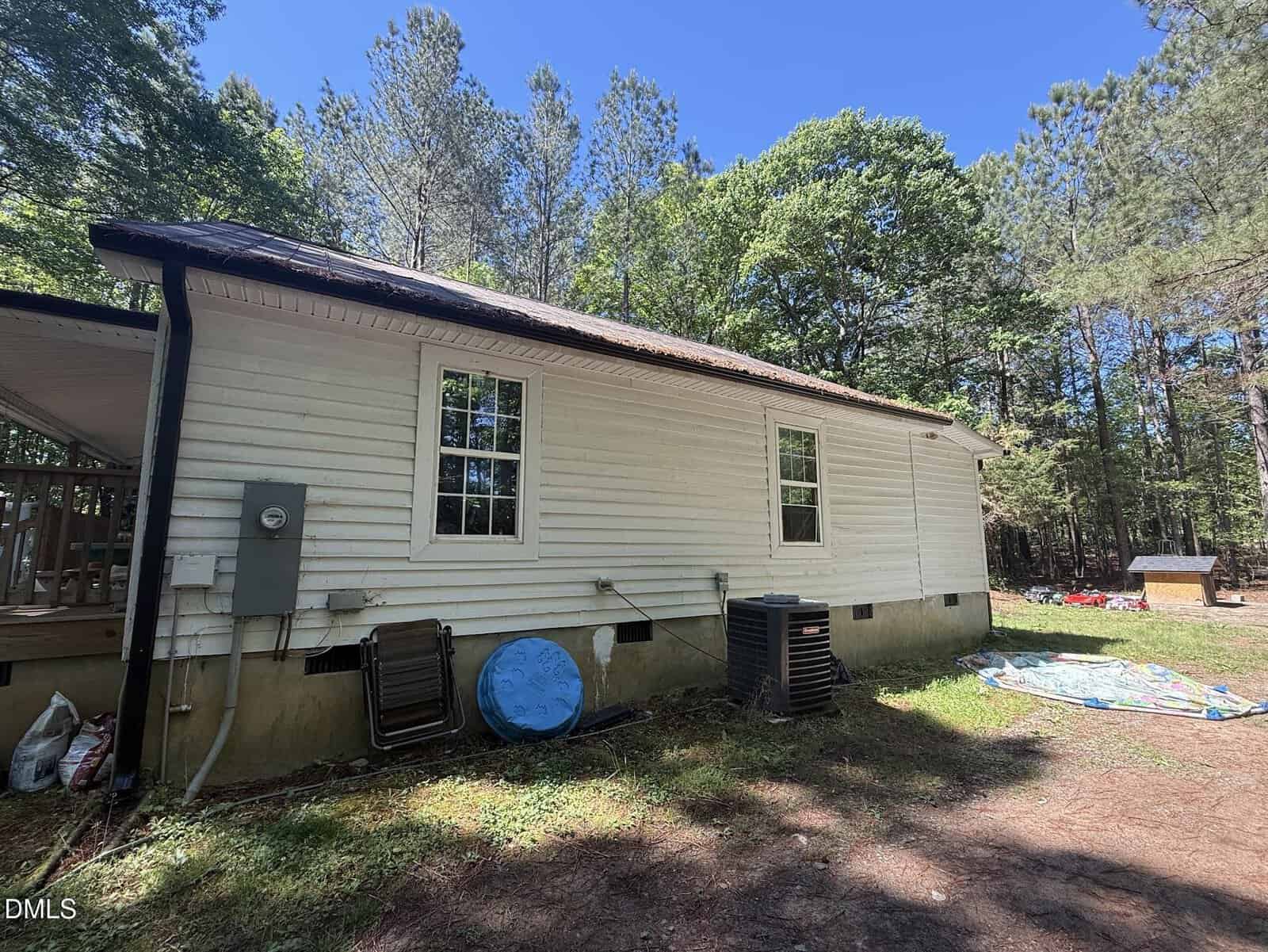 A white single-story house with two windows, HVAC units, and outdoor items on the ground beside it, surrounded by tall trees under a clear blue sky.