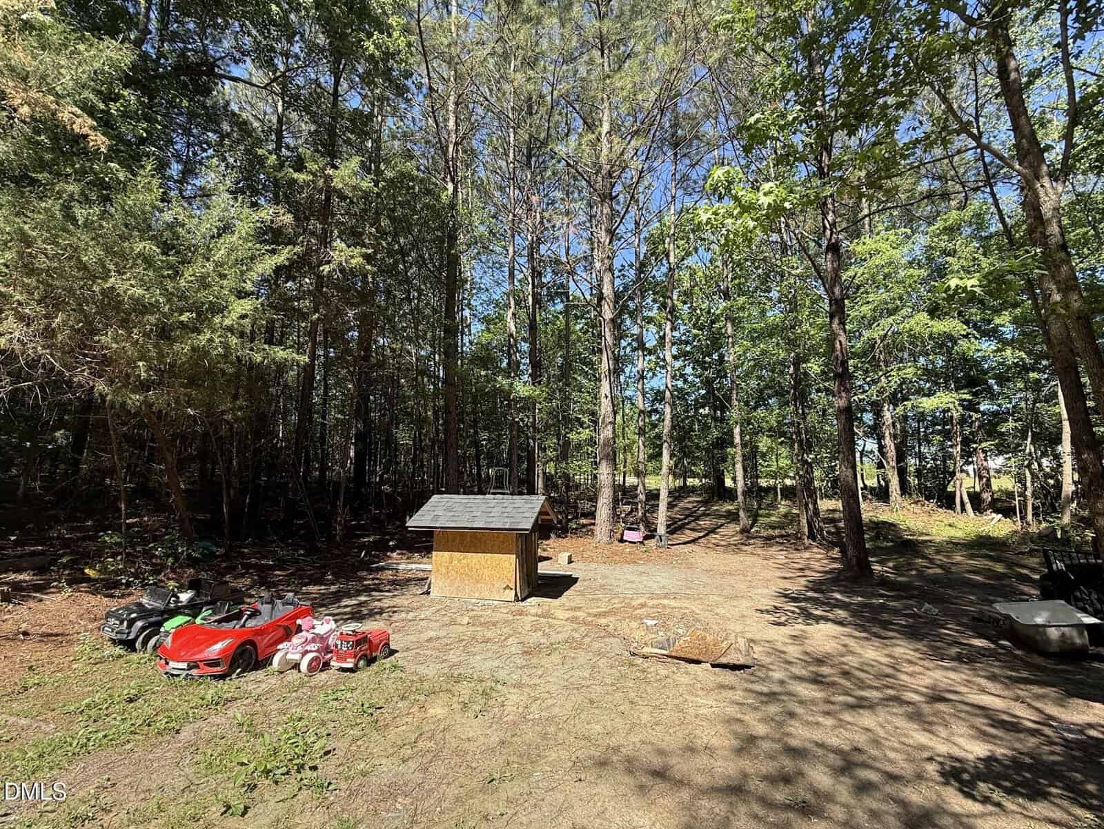A small wooden playhouse sits in a sunny clearing surrounded by tall pine trees. To the left are children's toy cars, including a red car, and to the right are scattered toys and a wheelbarrow. Bright blue sky overhead.