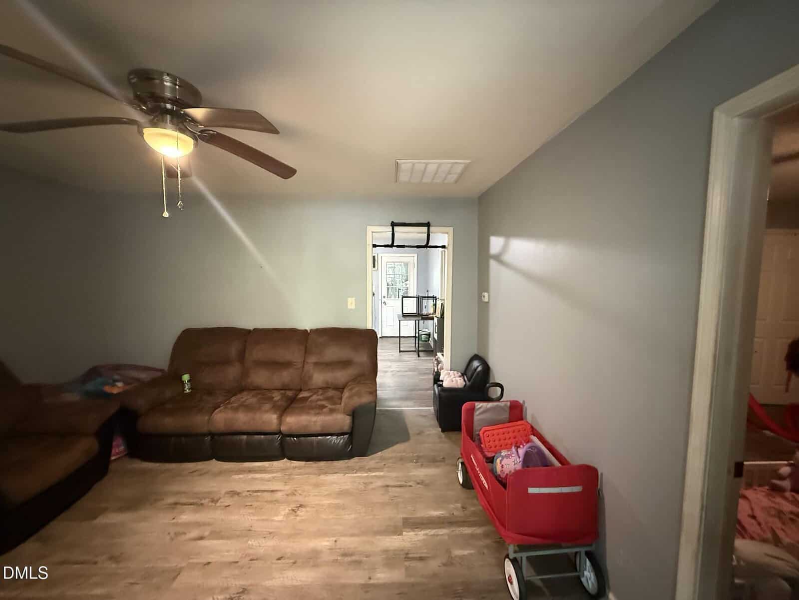 A living room with gray walls, brown sectional sofa, wood flooring, ceiling fan, and two red toy carts along the right wall. A doorway leads to another room with a table visible.