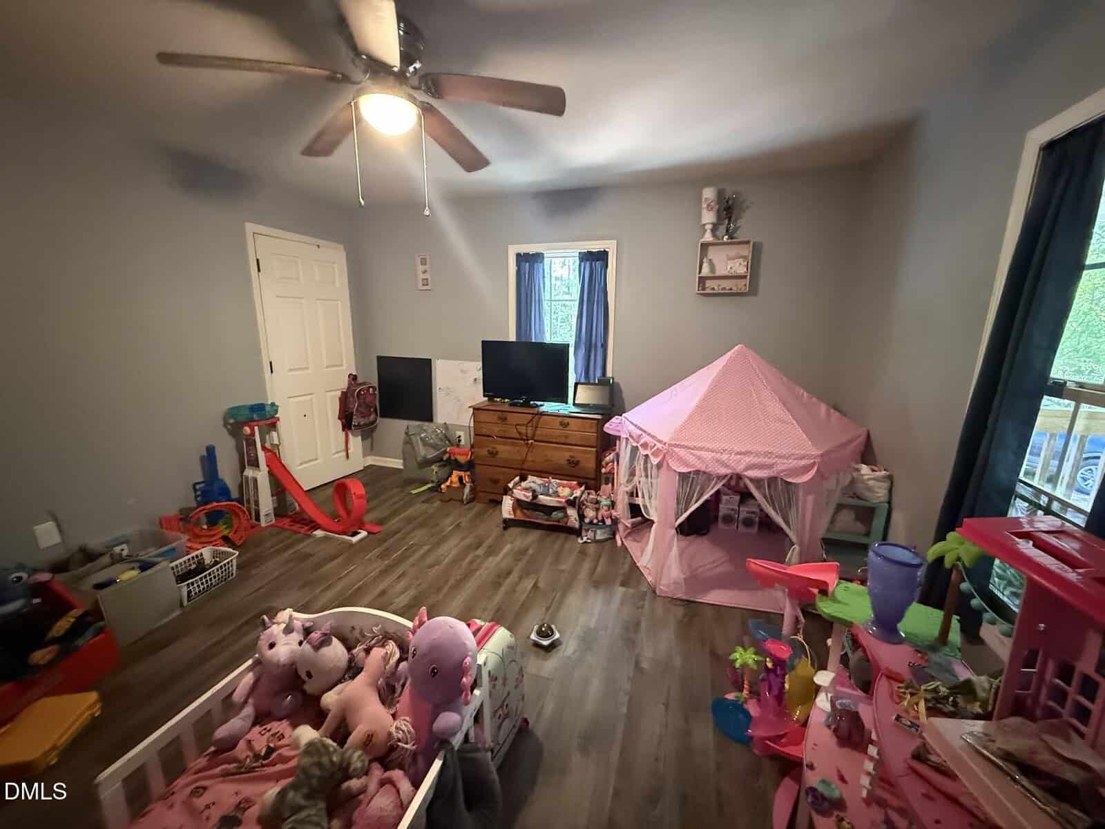 A child’s playroom with a bed full of stuffed animals, a pink tent, toys, a dresser with a TV, and a ceiling fan. The floor is wooden, and there are two windows with blue curtains letting in natural light.