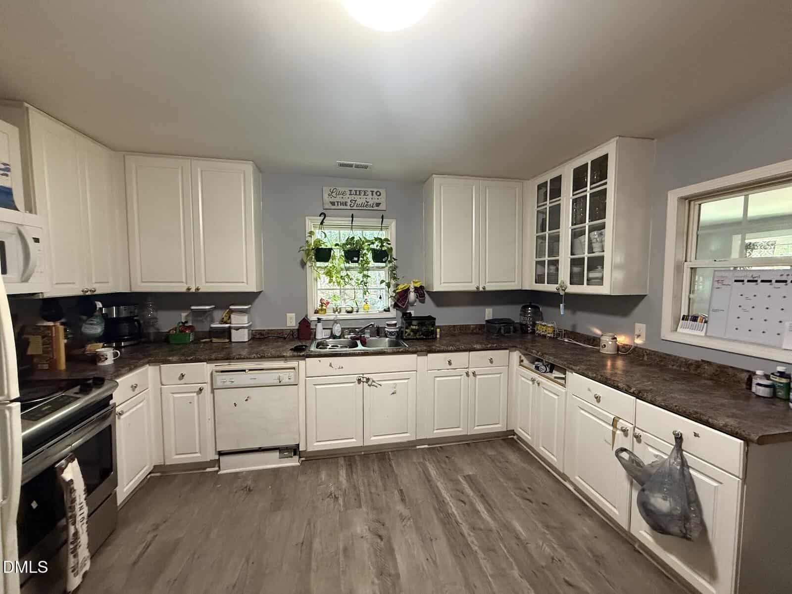 A cozy kitchen with white cabinets, brown countertops, a window above the sink with hanging plants, and appliances including a stove, dishwasher, and microwave. A calendar hangs by the window.