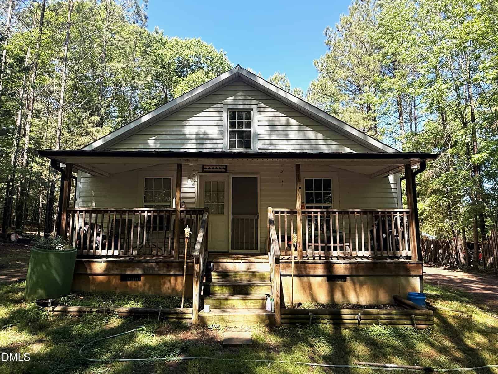 A small white house with a front porch and steps, surrounded by tall trees and greenery in a wooded area on a sunny day.