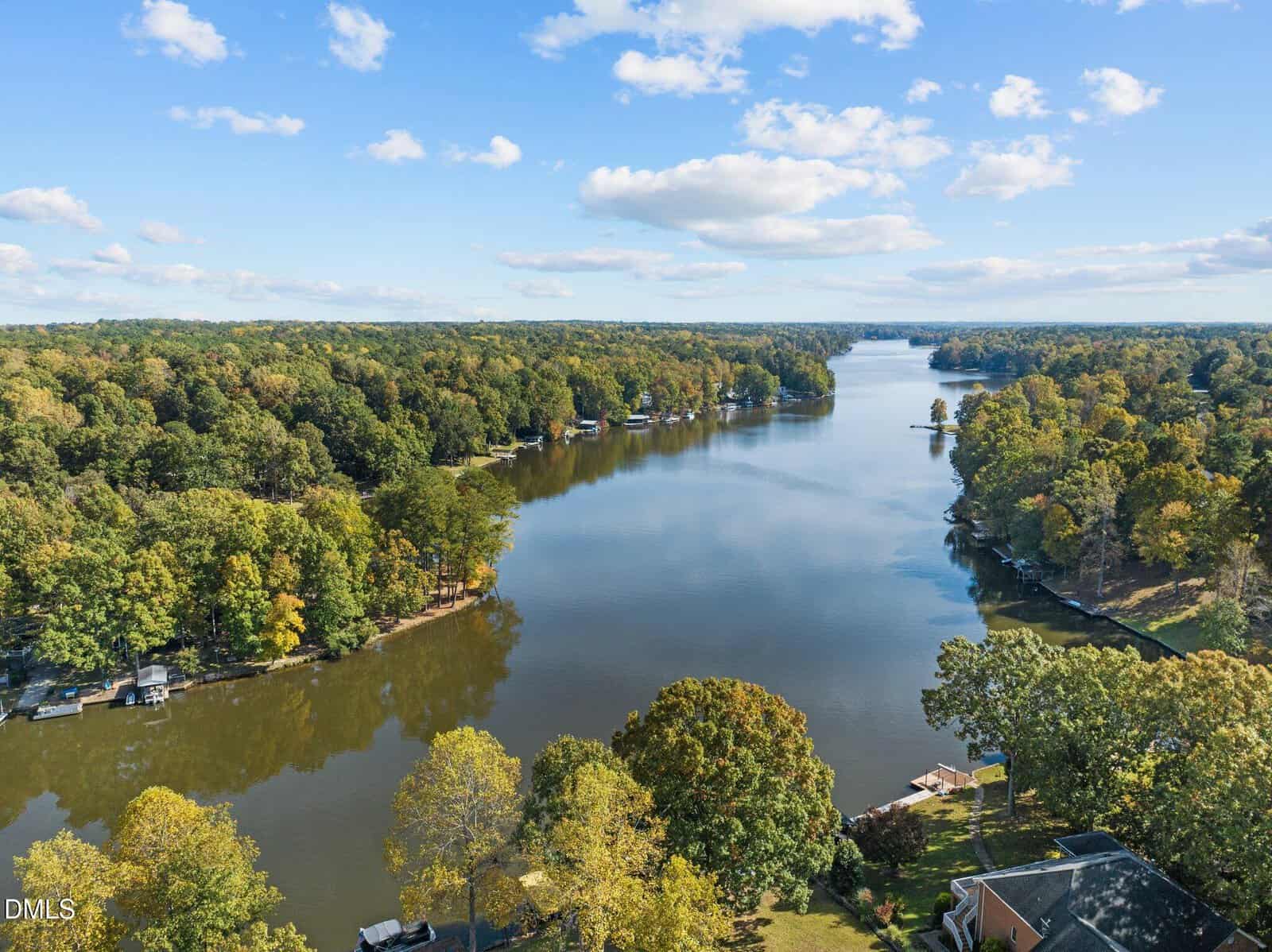 A wide river or lake surrounded by dense, green trees under a blue sky with scattered clouds; several houses and docks line the shoreline.