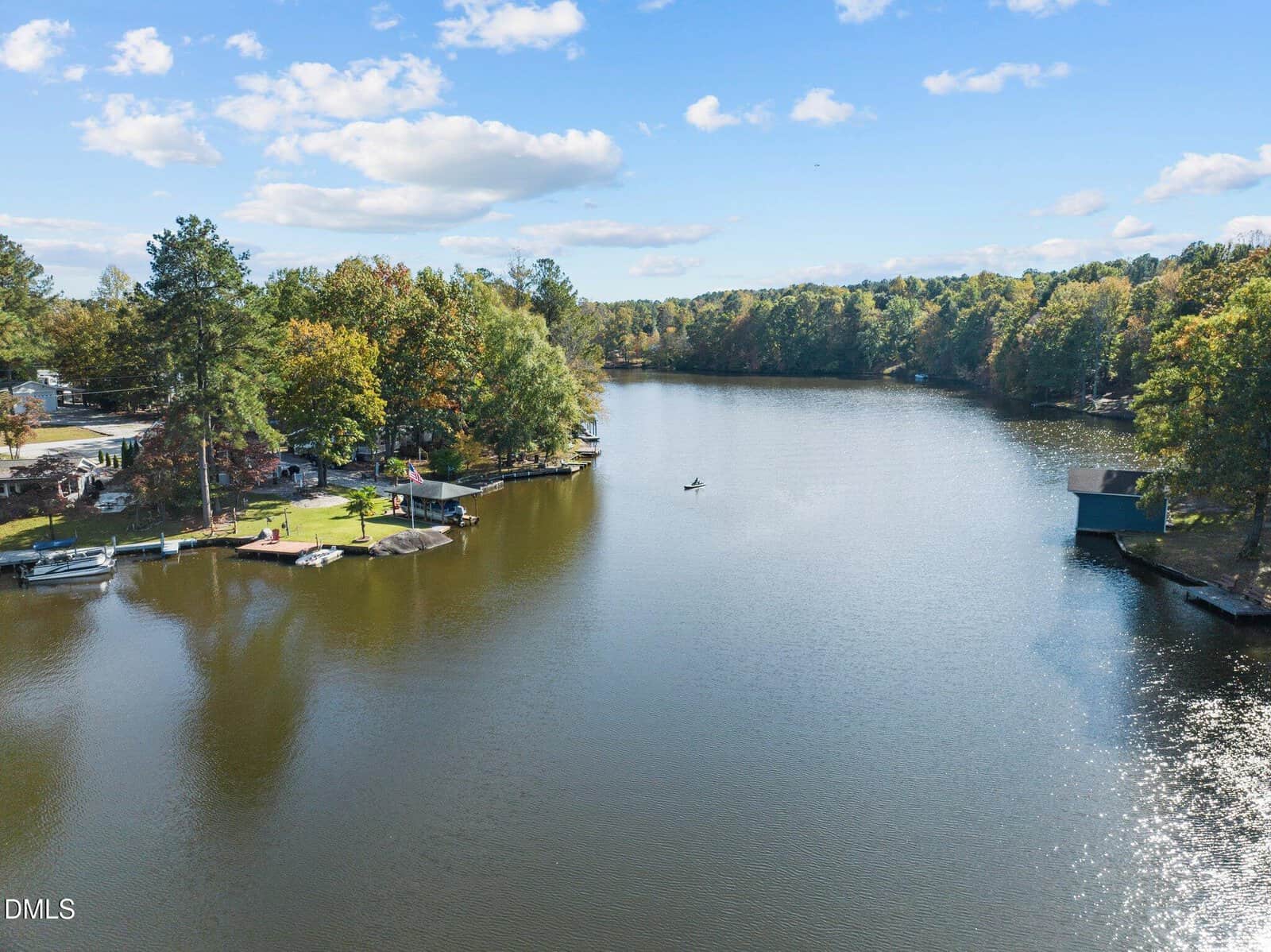 A calm lake surrounded by trees with docks and boats along the shoreline. A small boat is visible in the center of the water under a blue sky with scattered clouds.