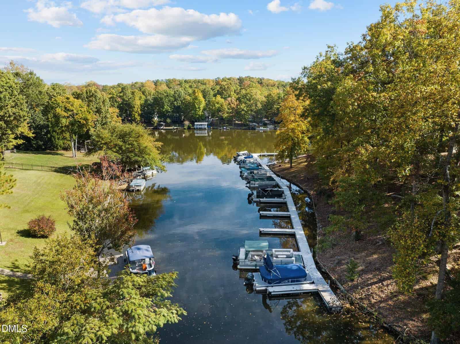 A peaceful lake with several boats docked along a wooden pier, surrounded by lush green trees and grassy areas on a sunny day with scattered clouds in the sky.