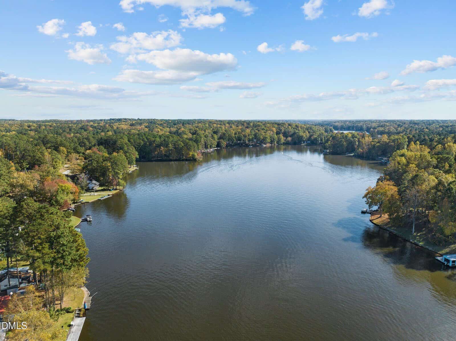 A wide, calm river is surrounded by dense, green and golden trees under a partly cloudy blue sky, with a few docks and boats along the shoreline.