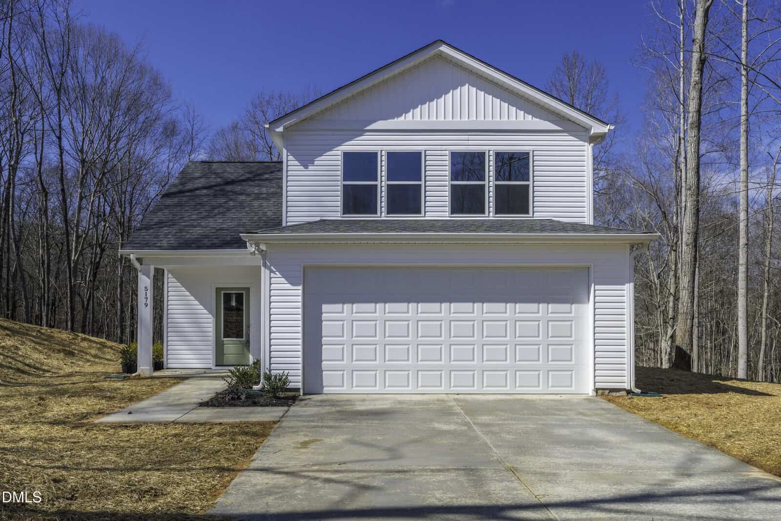 A two-story white house with gray roof shingles, attached two-car garage, concrete driveway, and single front door sits at 918 N Bickett Boulevard in Louisburg. Leafless trees frame the background under a clear blue sky and there is no grass on the front lawn.