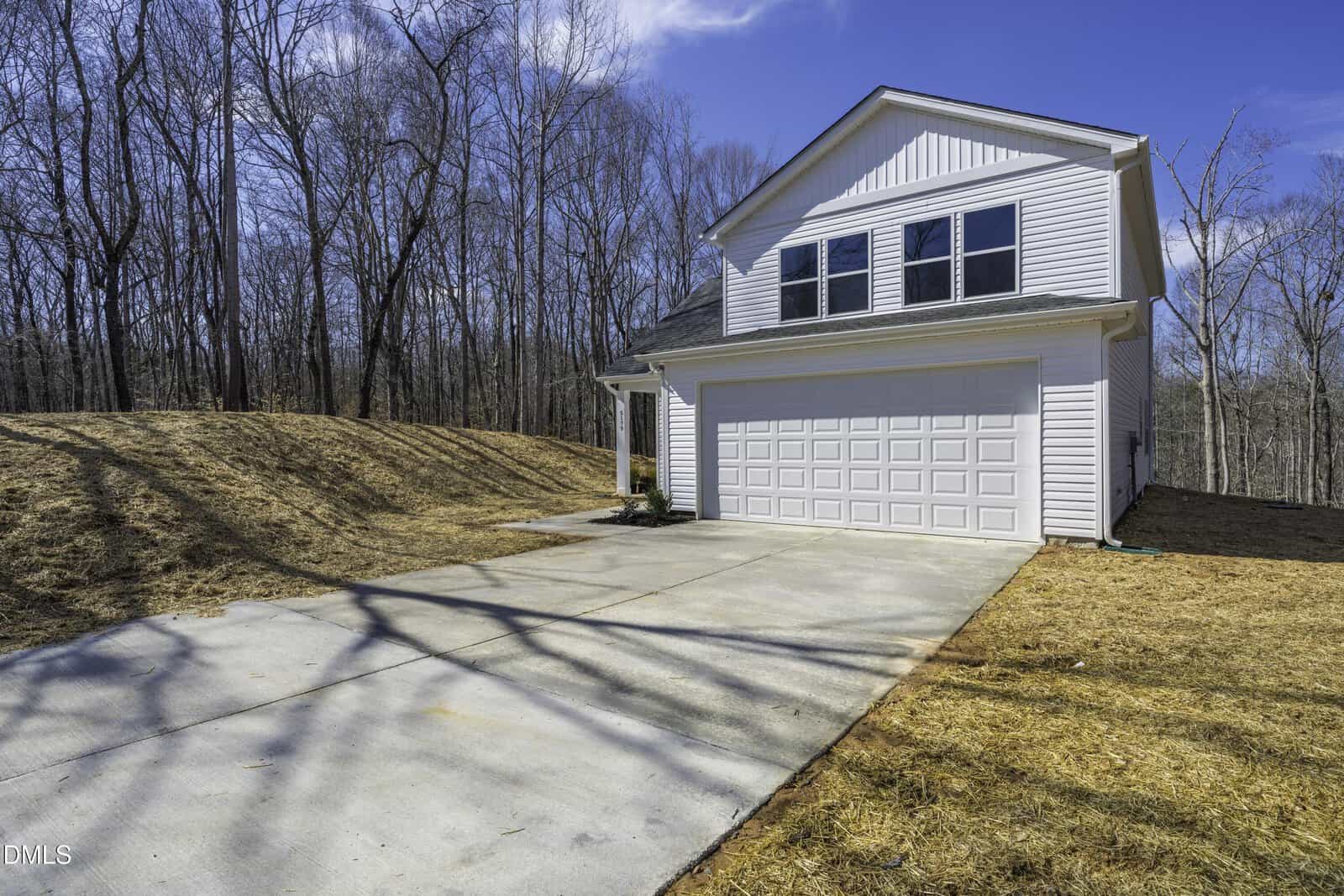 A two-story white house with a double garage sits beside a concrete driveway. The yard is covered in dry grass and surrounded by leafless trees under a clear blue sky.