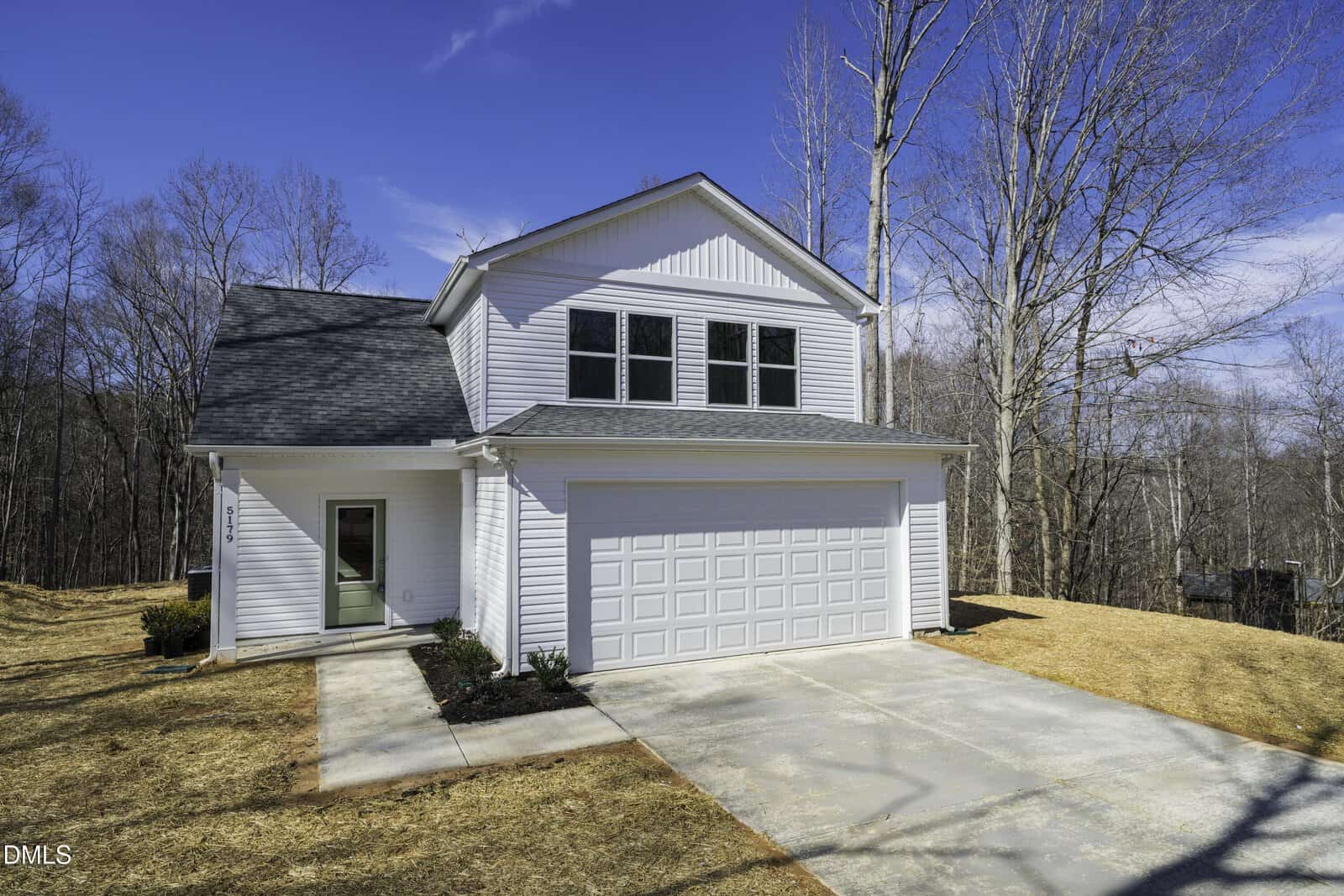Two-story white house with a double garage, front lawn with sparse grass, small landscaped area near the front door, and leafless trees in the background under a clear blue sky.