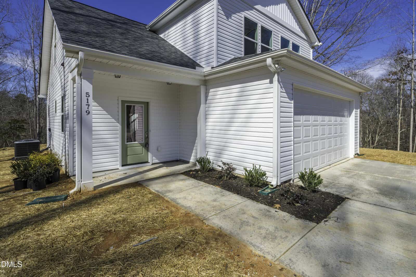 A white two-story house with a double garage, green front door, and covered entryway. There are small plants in a garden bed along the driveway and potted plants nearby. The grass is covered with straw.