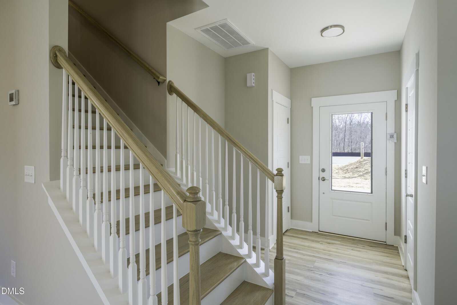 Bright entryway with a white front door, a window, light wood flooring, and a staircase with white spindles and wooden handrail. Neutral-colored walls and natural light create a welcoming atmosphere.