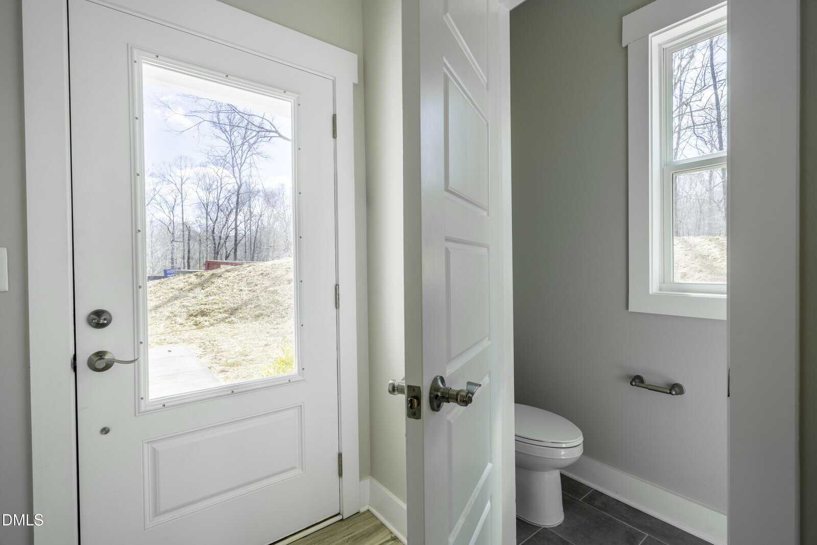 A white door with a window leads outside; next to it, an open bathroom door reveals a toilet and two windows with views of trees and a hill.