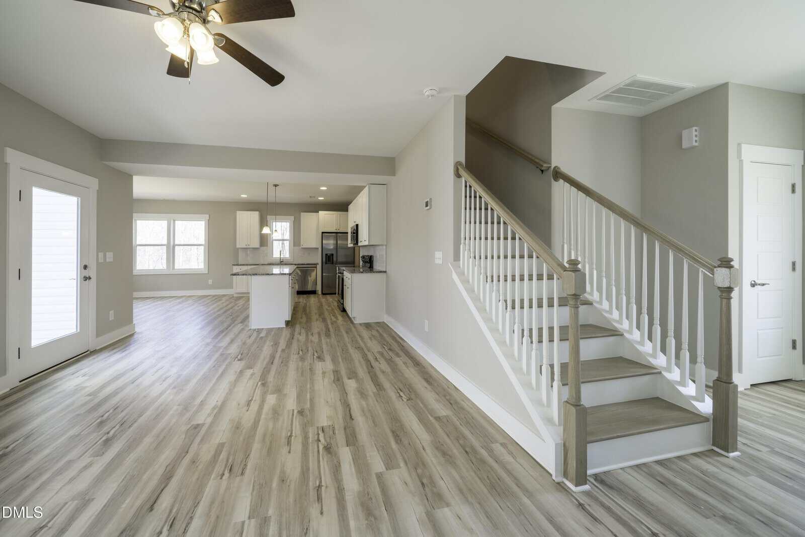 Open-plan home interior with light wood flooring, white walls, a staircase with white railing, and a modern kitchen with stainless steel appliances and an island. Ceiling fan and large windows provide natural light.