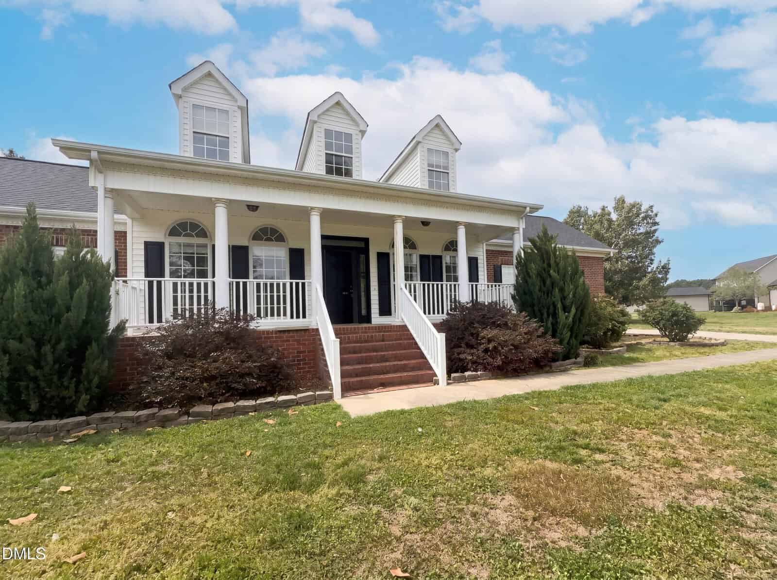 A two-story brick house with a large front porch, white columns, railings, and dormer windows. Brick steps lead to the entrance, surrounded by shrubs and a grassy lawn under a partly cloudy sky.
