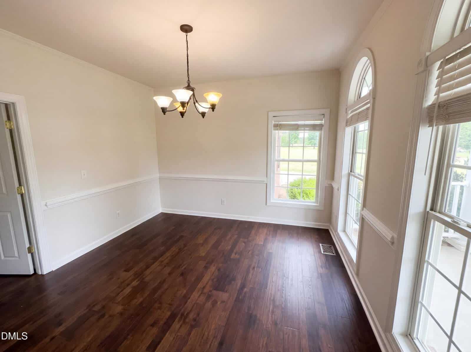A bright, empty room with hardwood floors, cream-colored walls, a chandelier, large windows with blinds, and white trim. Natural light fills the space, and a door is visible on the left.
