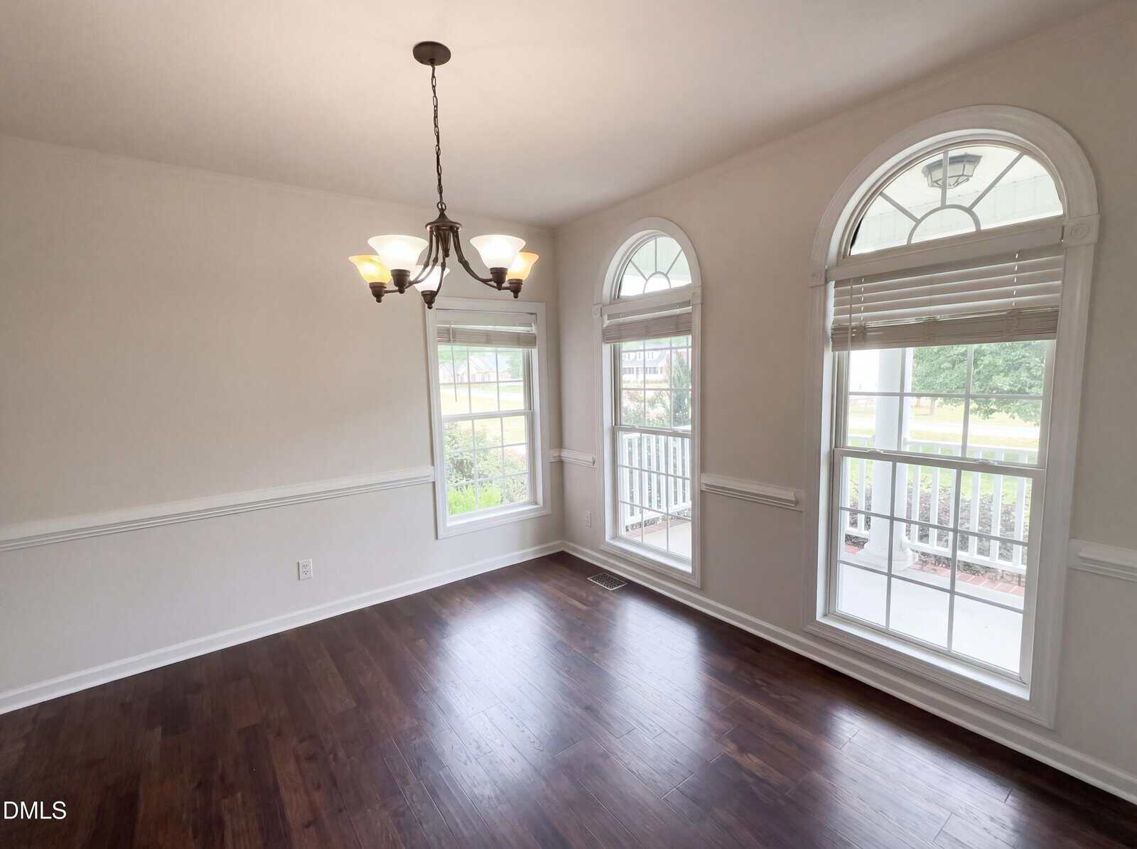 Bright dining room with dark wood floors, three large windows with white trim and blinds, white walls, and a black chandelier hanging from the ceiling. The view outside shows greenery and a front porch.