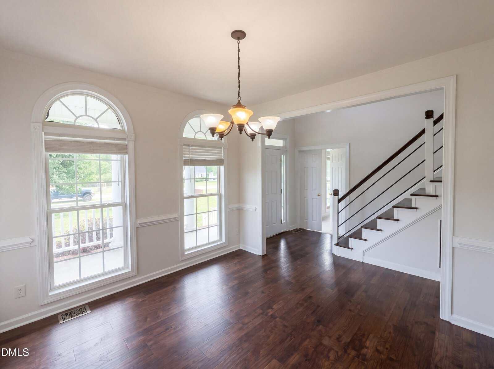 A bright room with dark wood floors, three large arched windows, white walls, a modern chandelier, and an open entryway with stairs featuring a black railing. Natural light fills the space.