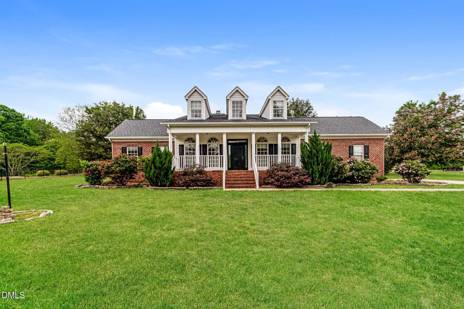 A red brick house with white trim, three dormer windows, and a large front porch with columns, surrounded by green lawn and landscaped bushes, under a clear blue sky.