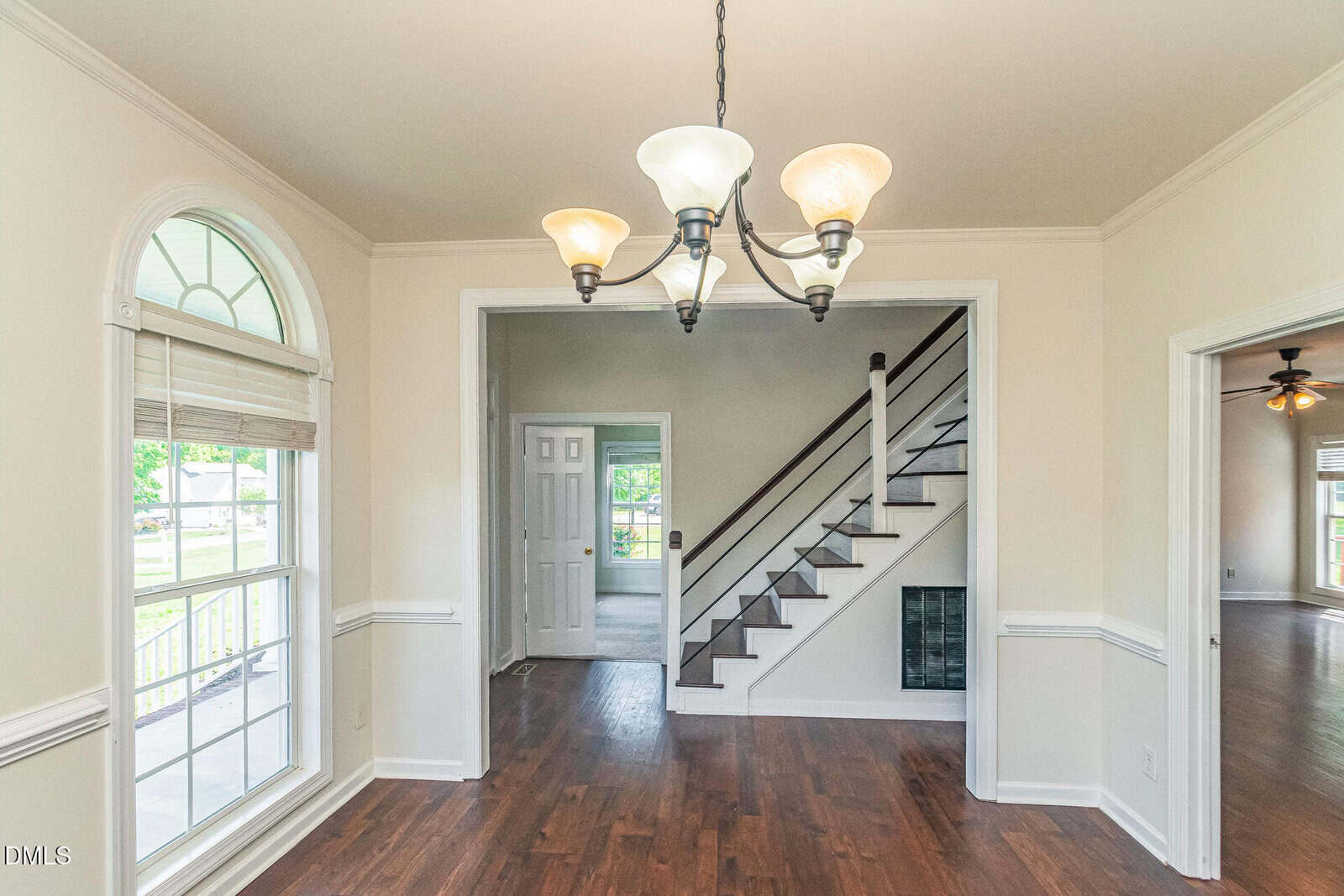 Bright dining room with hardwood floors, white walls, a chandelier, large window with blinds, and a view into a hallway with stairs and another room with a ceiling fan.