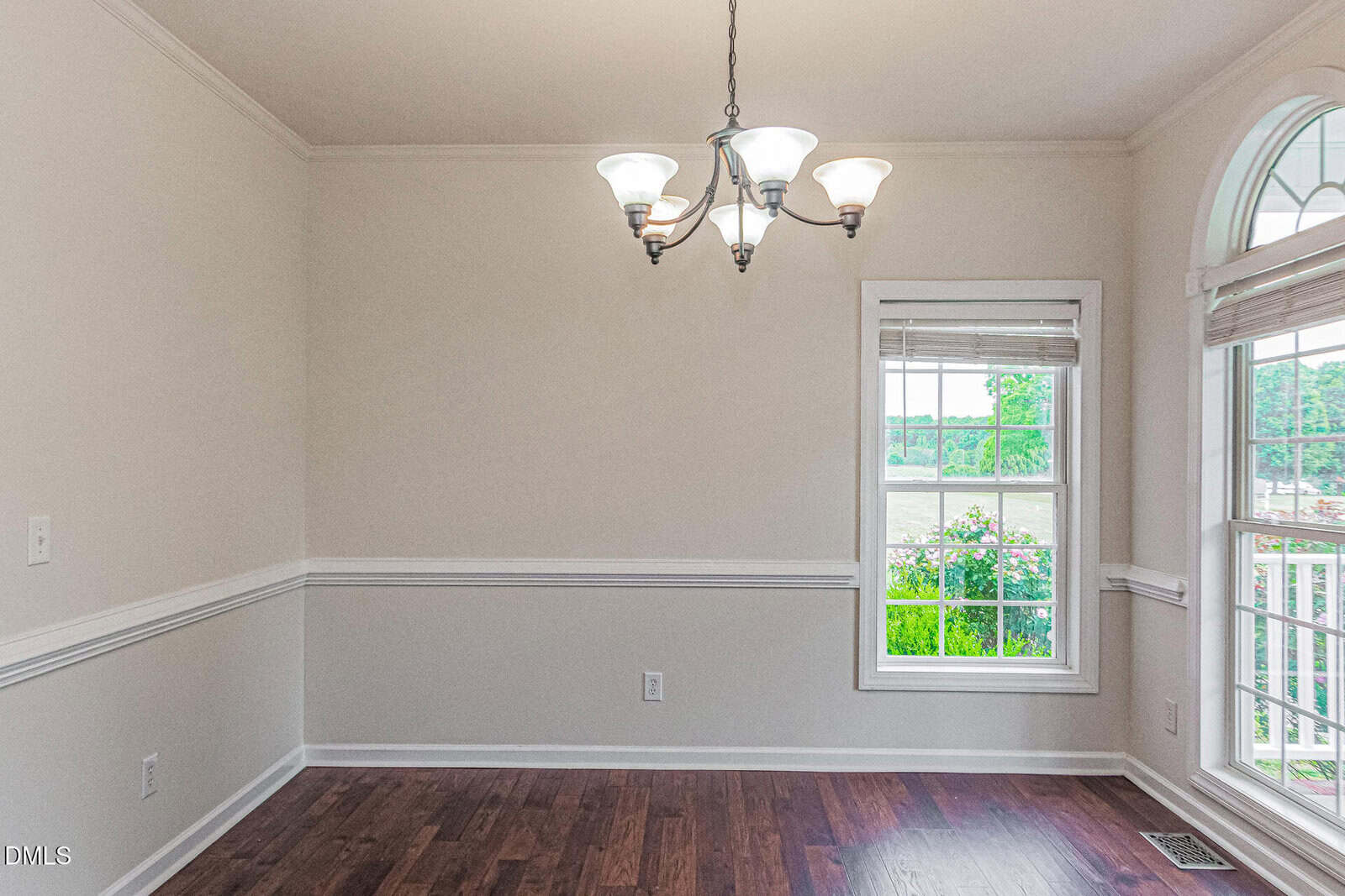 A bright, empty dining room with hardwood floors, white walls, a window showing greenery outside, and a chandelier hanging from the ceiling.