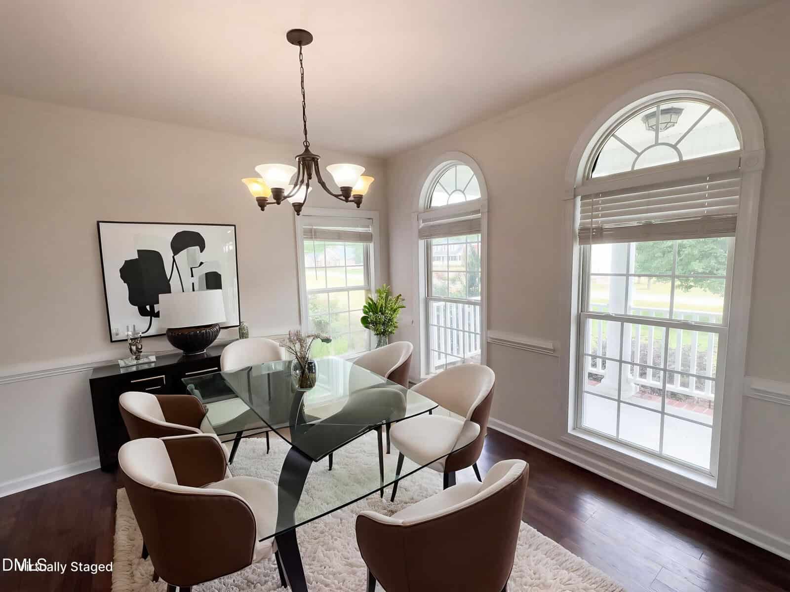 A bright dining room with a glass table, six beige chairs, a light fixture above, large arched windows with blinds, and modern black-and-white artwork on the wall. A vase with flowers sits on the table.