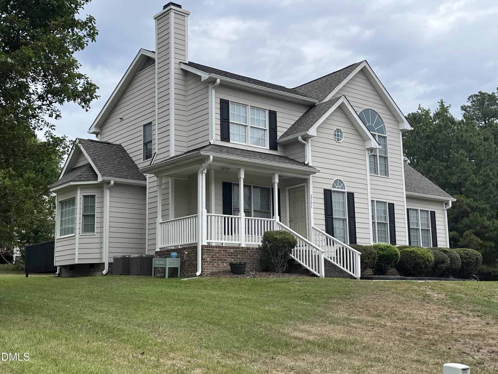 A two-story beige house with white trim, dark shutters, a covered front porch, brick foundation, and multiple large windows, surrounded by green grass, shrubs, and trees under a cloudy sky.