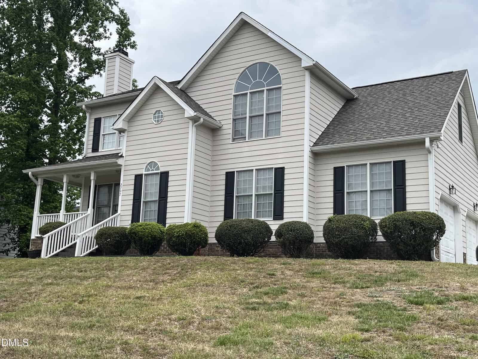 A two-story beige house with white trim and dark shutters, featuring a covered front porch, arched windows, and neat shrubs in front on a sloped lawn.