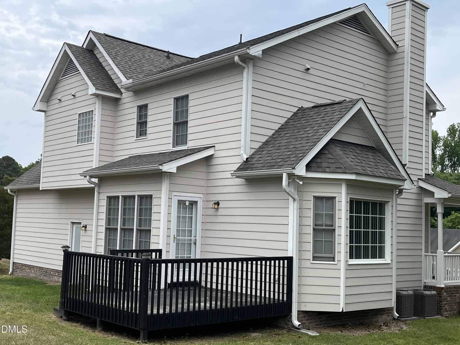 Two-story beige house with white trim, multiple windows, a black-railed wooden deck, and a small bay window section, set against a grassy yard and trees.
