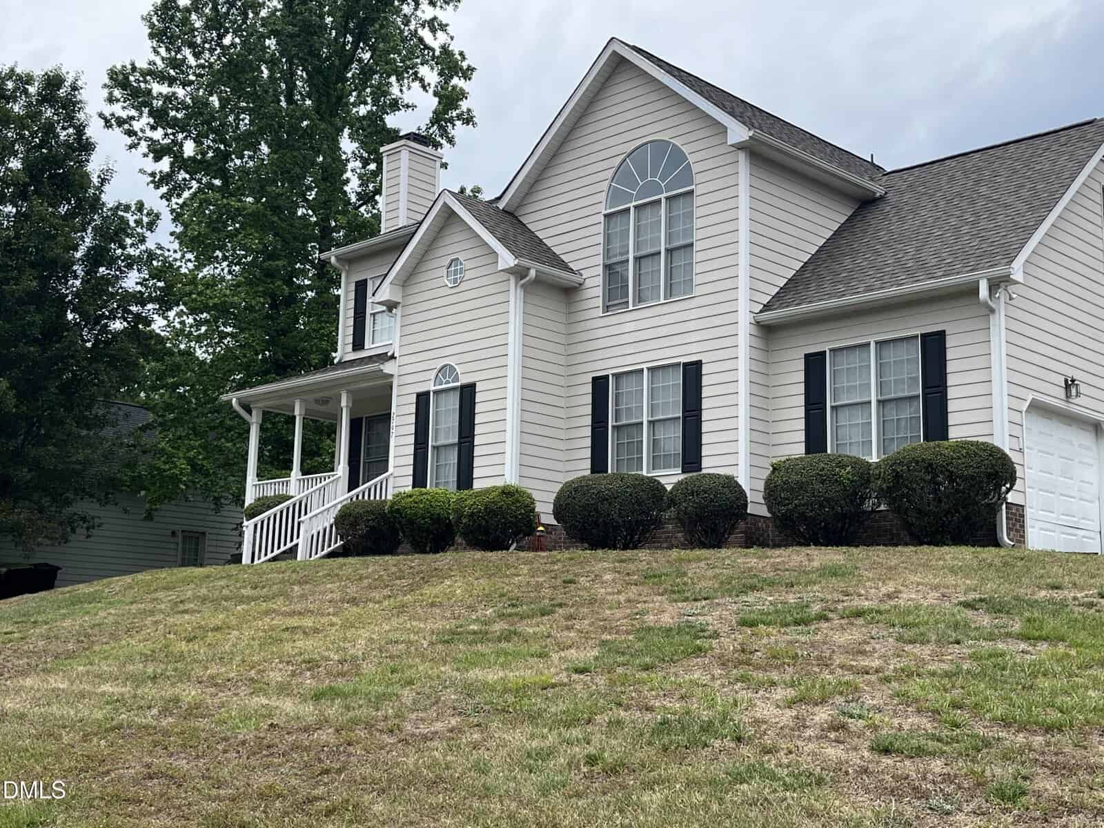 A two-story beige house with black shutters, white trim, a front porch, manicured bushes, and a two-car garage sits on a sloped lawn with patchy grass and trees in the background.